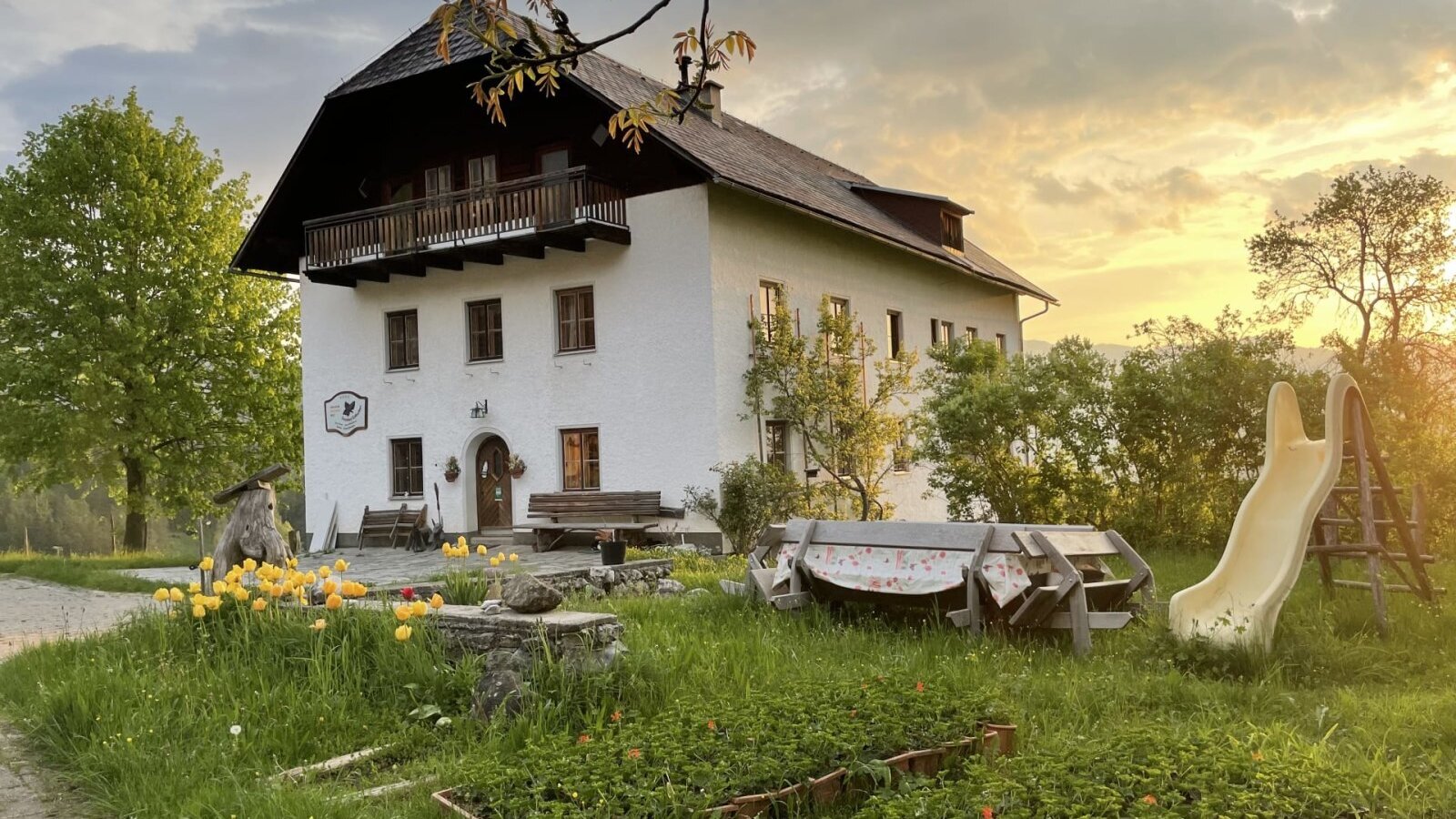 The farmhouse with a balcony, surrounded by a garden with a slide and outdoor seating at sunset.