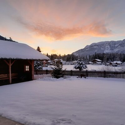 Winter landscape of the farmhouse with a snow-covered garden, wooden gazebo, and mountains in the background.
