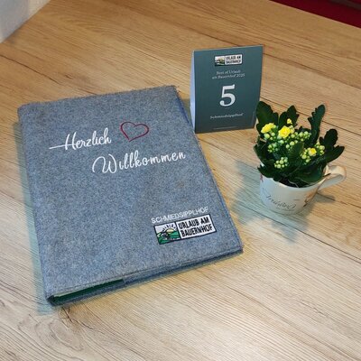A welcome binder from Schmiedsipplhof Urlaub am Bauernhof, an information card, and a small potted plant are displayed on a wooden table at the farmhouse.