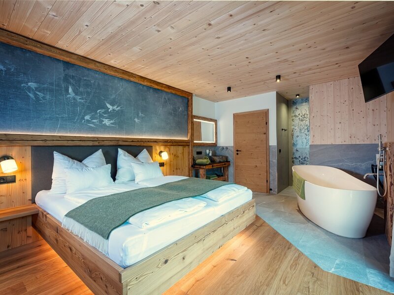 Double bed in the chalet with a freestanding bathtub, wooden elements, and a wall-mounted TV.