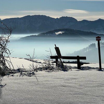 A snowy mountain landscape from a hiking path near the farm house, featuring a wooden bench and a trail marker.