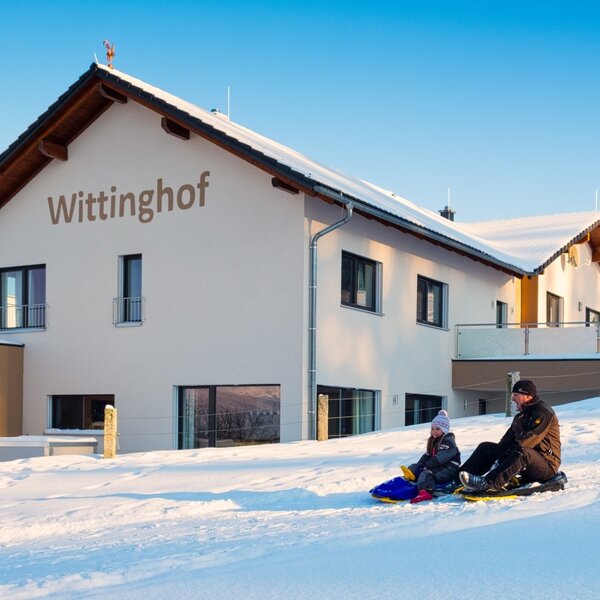 The Wittinghof Farm House exterior in winter, featuring a man and child sledding on the snowy hill adjacent to the property.
