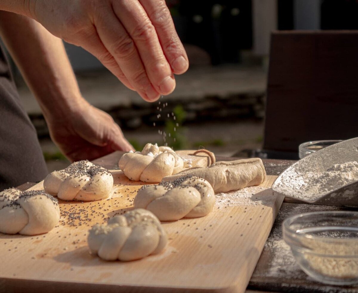 Hand sprinkling seeds on unbaked bread rolls on a wooden board, highlighting baking activities at the Bauernhof.
