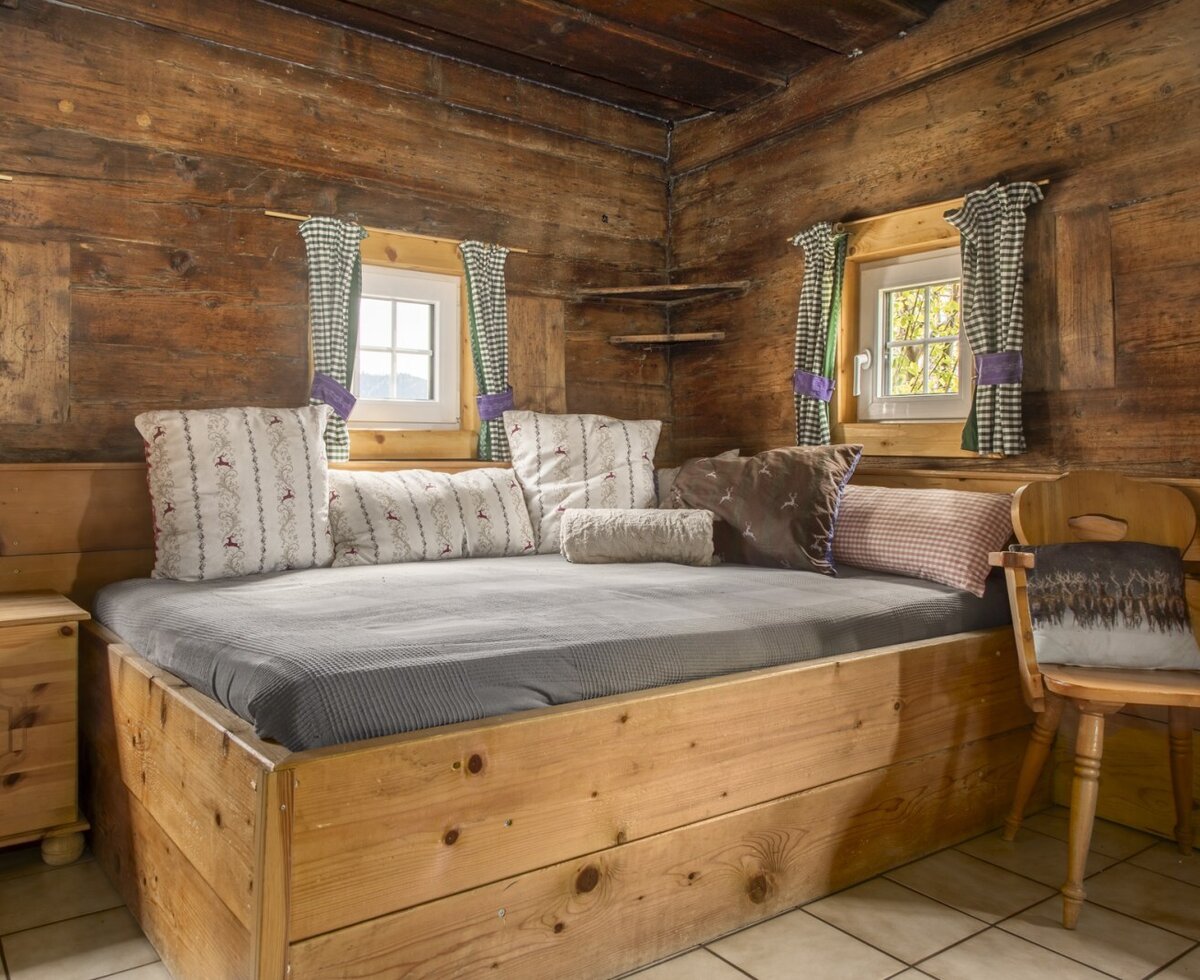 Bedroom in the farmhouse featuring a wooden daybed with cushions, wooden paneled walls, and windows with checked curtains.