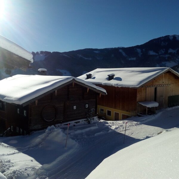 The Almhütte exterior with snow-covered roofs and grounds, surrounded by mountains on a clear, sunny day.
