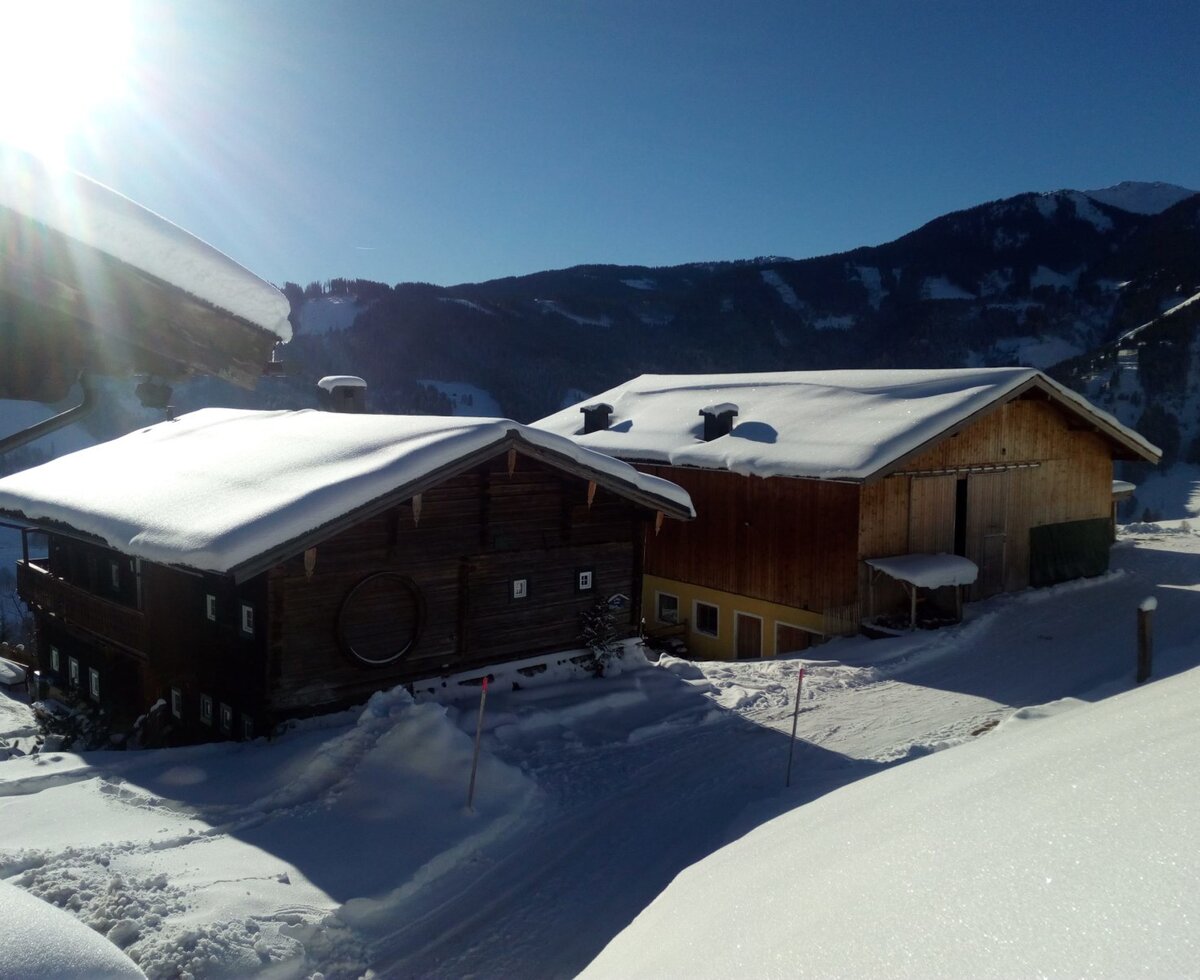 The Almhütte exterior with snow-covered roofs and grounds, surrounded by mountains on a clear, sunny day.