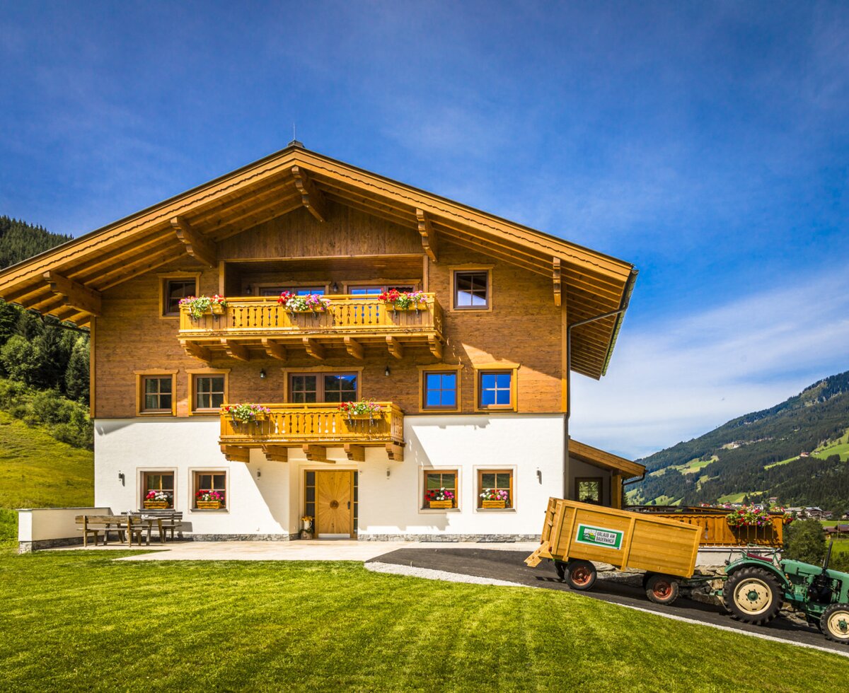 The farm house exterior features wooden balconies with flower boxes and an outdoor seating area, with a tractor on the driveway and mountains in the background.