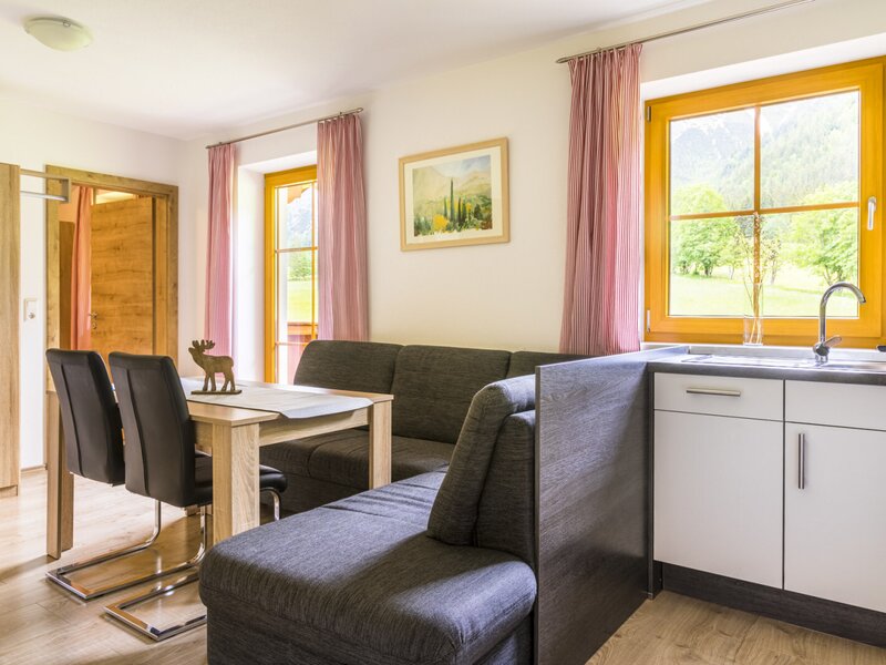 Living and dining area in an apartment of the farm house, featuring a corner sofa, a wooden dining table, and a kitchenette with a sink, all with windows offering views of the surrounding area.