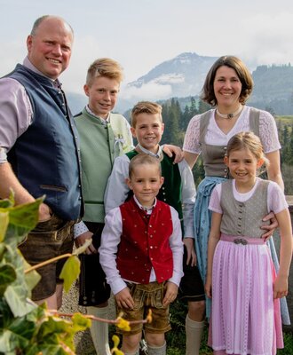 The hosts of the Farm House, a family in traditional attire, are pictured outdoors with a mountain backdrop.