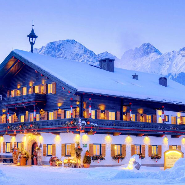 The traditional Farm House exterior with illuminated windows, balconies, and a snow-covered roof, set against a backdrop of snowy mountains.