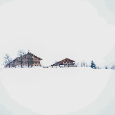 Two wooden farmhouses stand on a snow-covered hill surrounded by trees.