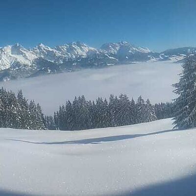 Panoramic view of snow-covered mountains, pine trees, and a cloud-filled valley in the winter landscape surrounding the farmhouse.