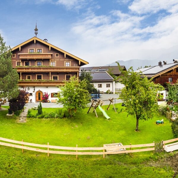 An inviting alpine farmhouse surrounded by a spacious green lawn, featuring a children's play area and grazing cows in the background.
