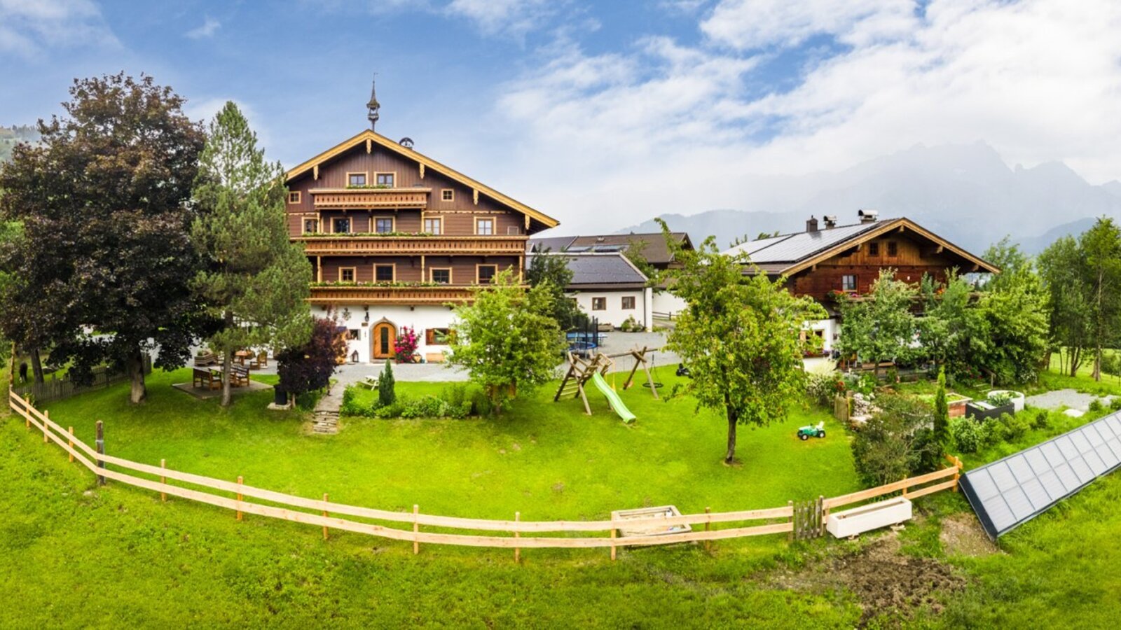 An inviting alpine farmhouse surrounded by a spacious green lawn, featuring a children's play area and grazing cows in the background.