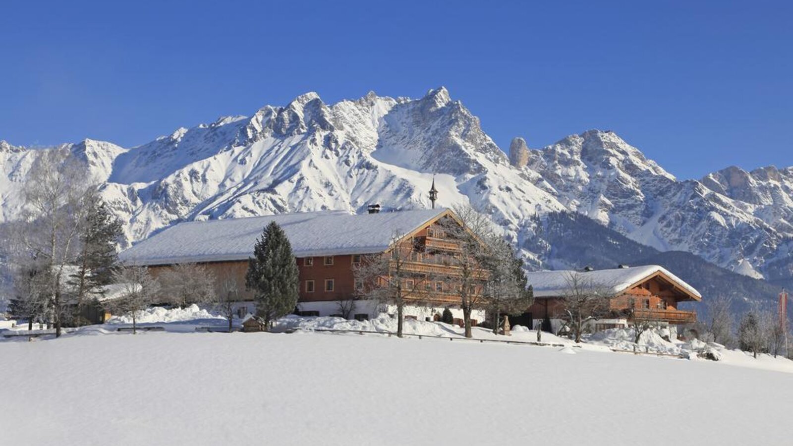 The traditional wooden Farm House stands in a snow-covered landscape with prominent snow-capped mountains in the background.