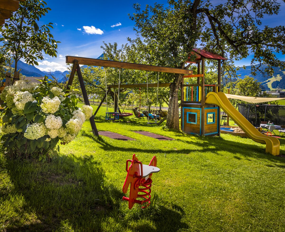 The farm house garden features a children's playground with a swing set, slide, sandpit, trampoline, and hammocks, surrounded by trees and mountain views.