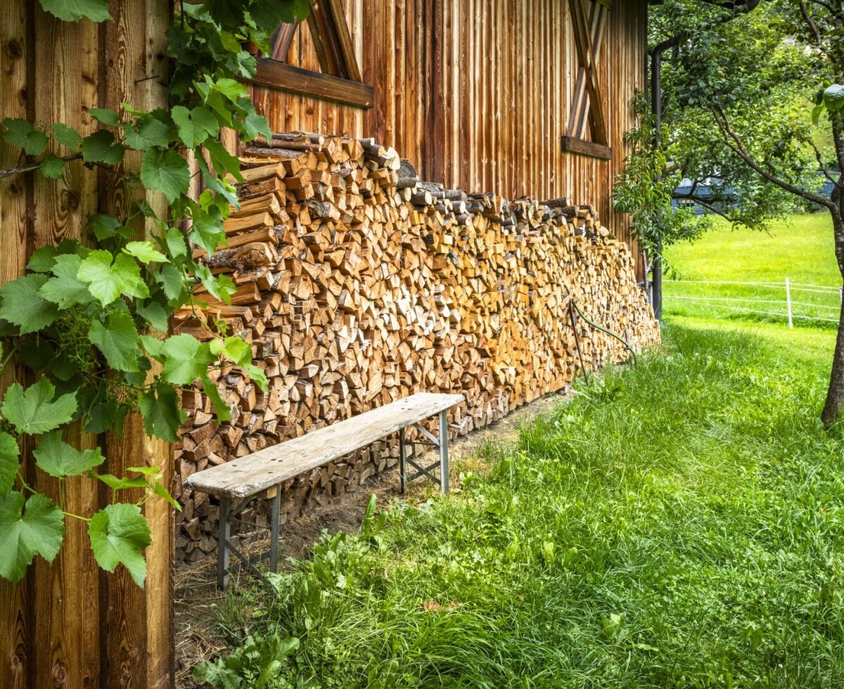 An idyllic outdoor spot at the farmhouse, featuring a rustic wooden bench set against neatly stacked firewood and a lush green lawn.