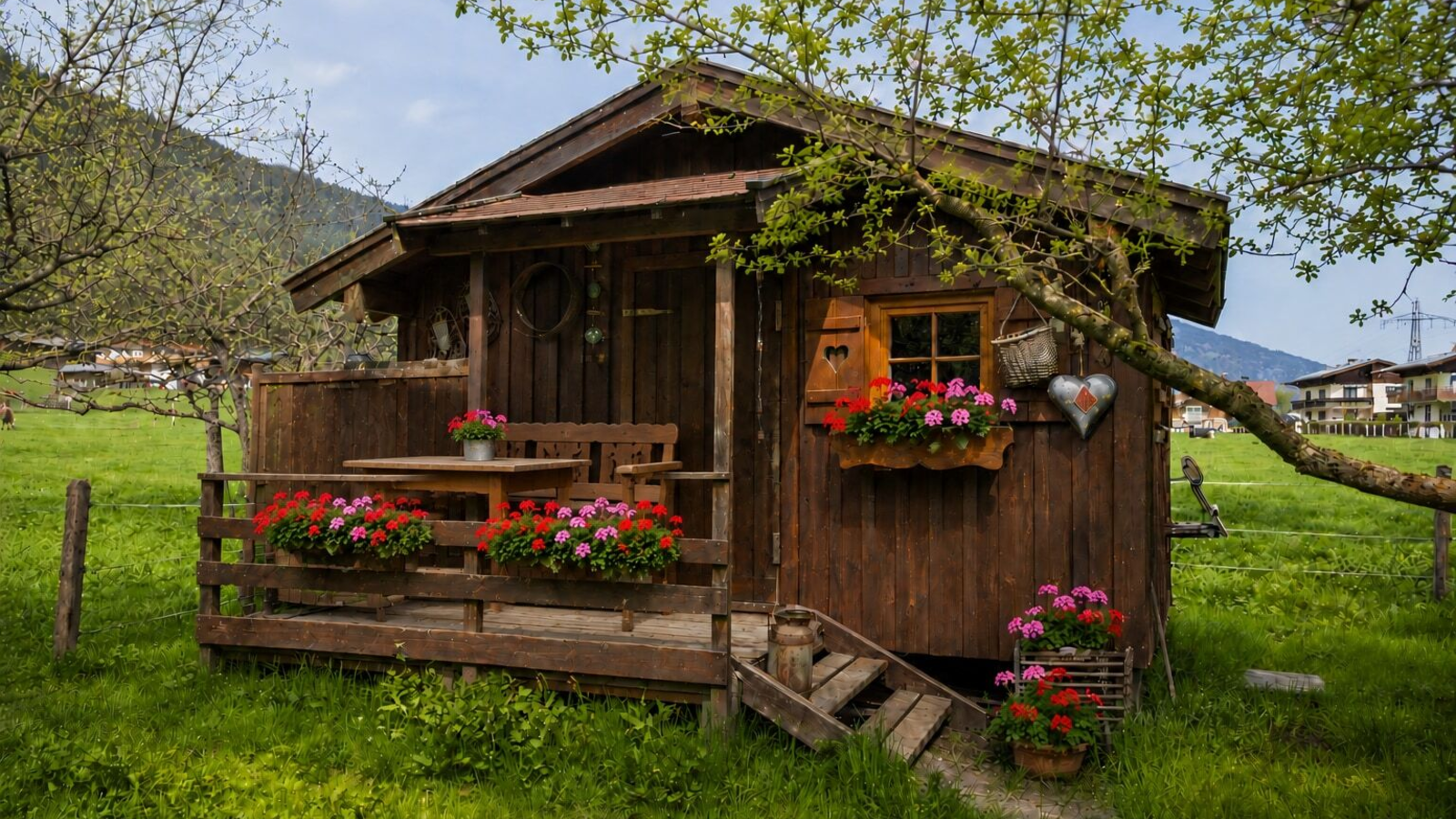 Wooden garden hut with a small terrace, featuring a wooden table, benches, and flower boxes, set in a green lawn.