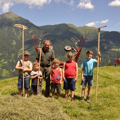 An adult and children holding rakes during farm work at the Farm House, set against a mountain backdrop.