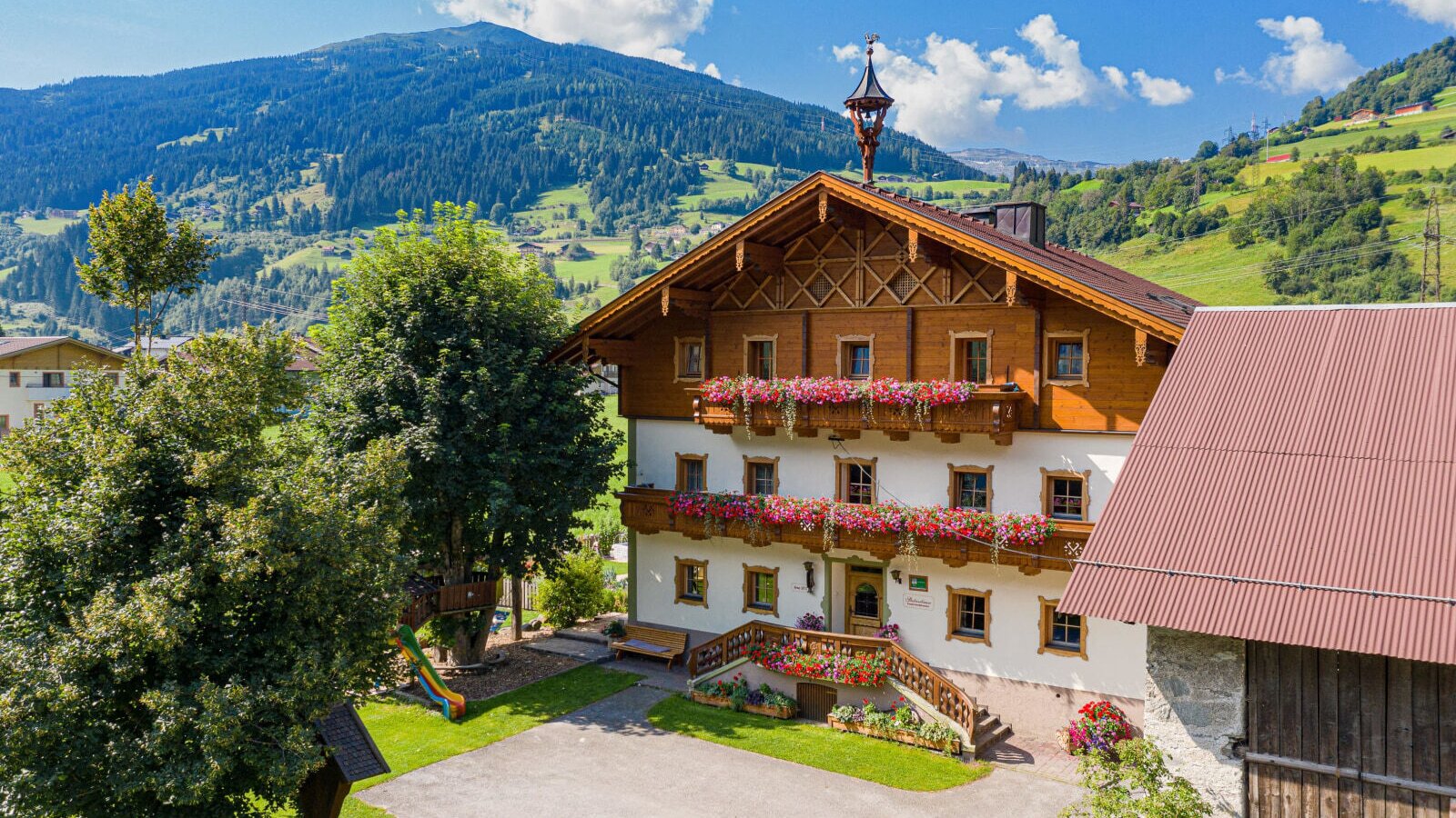 The farmhouse exterior, featuring wooden balconies with flowers, a green lawn with trees, a children's slide, and a mountainous backdrop.