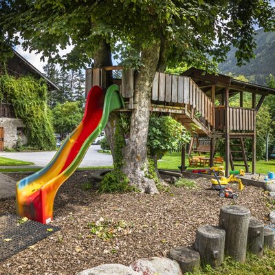The farmhouse's inviting children's playground features a colorful slide, a treehouse, and a swing set, set against a scenic mountain backdrop.