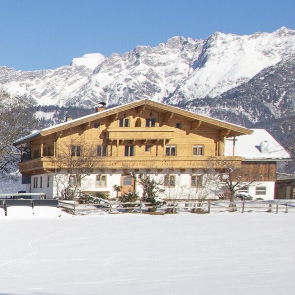 The exterior of the Farm House, featuring a wooden facade, balconies, and a snow-covered landscape with mountains in the background.