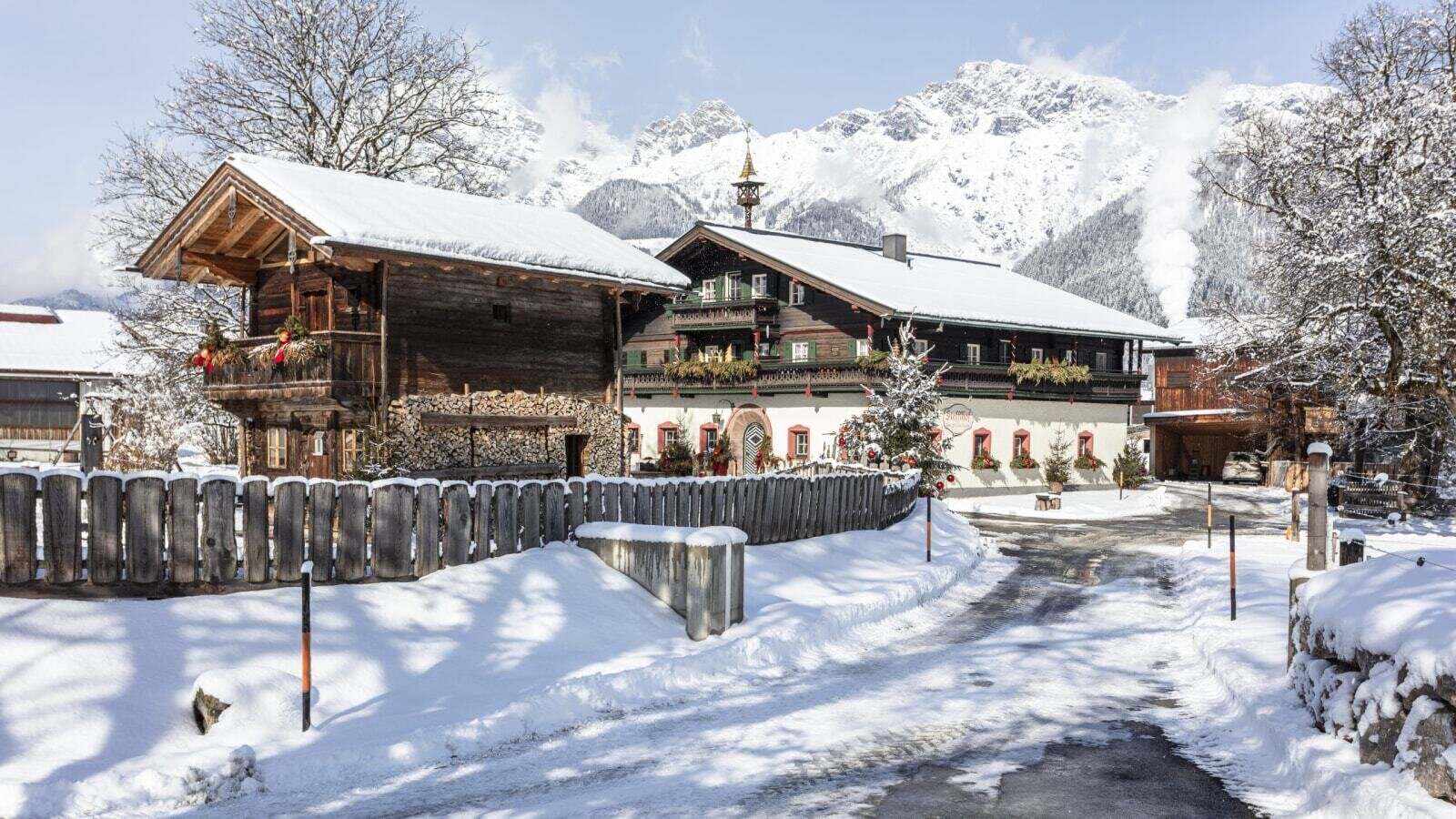 Exterior view of the farmhouse in winter, featuring snow-covered roofs, traditional wooden construction, and mountains in the background.