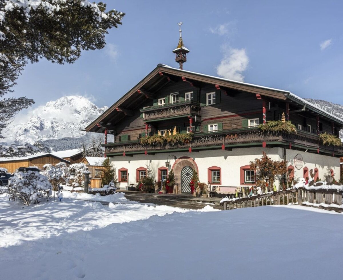 The farmhouse in winter with a snow-covered forecourt, balconies, a decorated Christmas tree, and mountain backdrop.