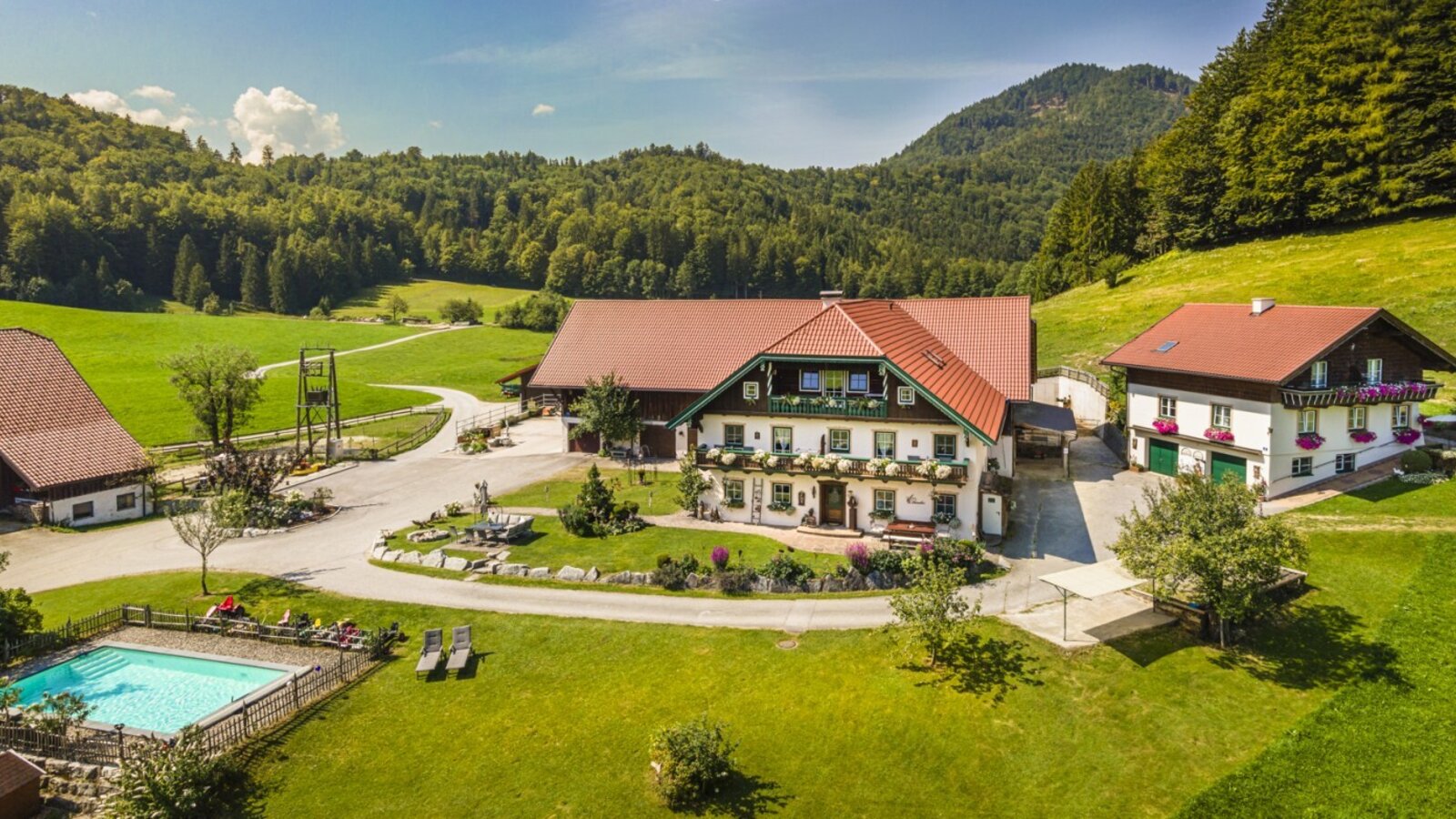 The farm featuring multiple buildings, an outdoor swimming pool, and expansive green areas, set against a backdrop of forests and mountains.