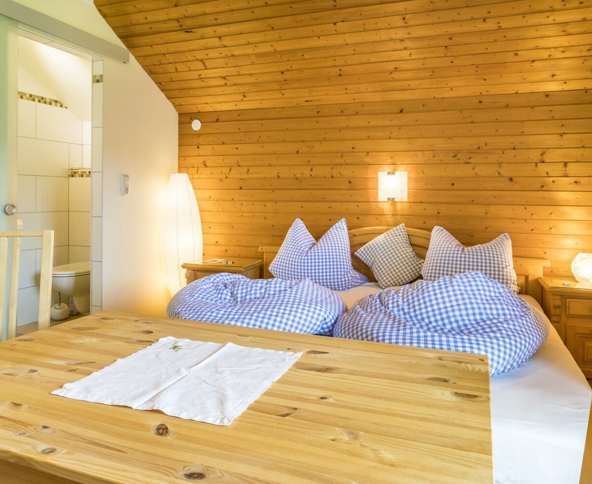 Bedroom area of the Ferienwohnung with a double bed, blue and white checkered bedding, wooden walls, a dining table, and an adjacent bathroom.