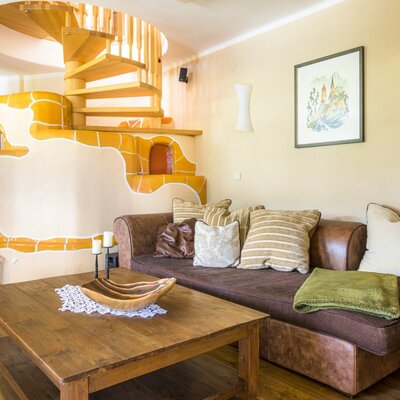 The living area of the farm house features a brown sofa, a wooden coffee table, a tiled stove, and a wooden spiral staircase.