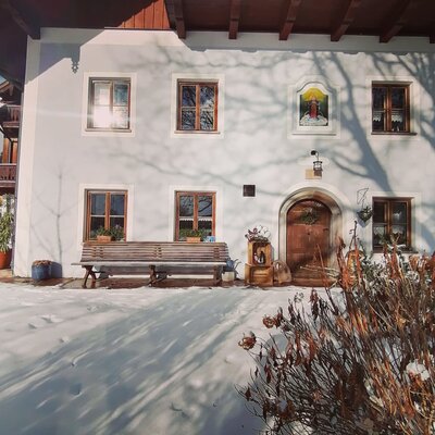 The Farm House exterior, featuring white walls, wooden windows, a wooden balcony, and a bench, with snow on the ground.