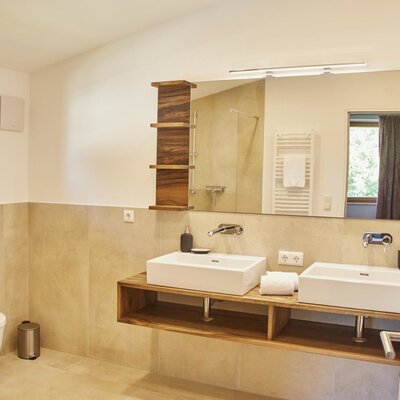 The bathroom in the Farm House features a double vanity with two sinks set on a wooden counter, a large mirror with integrated lighting, a toilet, and a shower with a glass partition, with a window providing natural light.