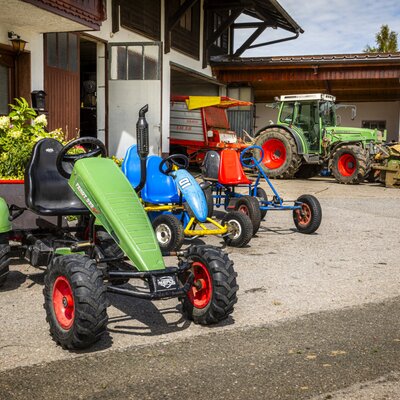 The farmhouse's inviting courtyard featuring colorful pedal go-karts for children and agricultural tractors in the background.