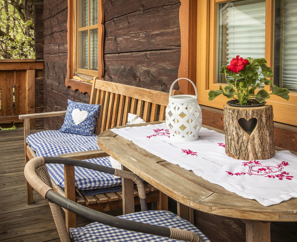 Farmhouse balcony with a wooden table, bench, checkered cushions, and a red geranium.