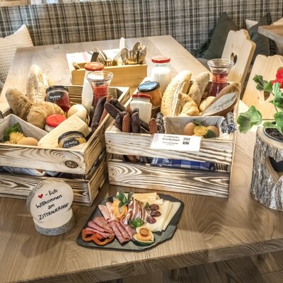 A rich farmhouse breakfast featuring fresh baked goods, cold cuts, cheese, jams, and drinks, presented in wooden baskets on a table at the farmhouse.