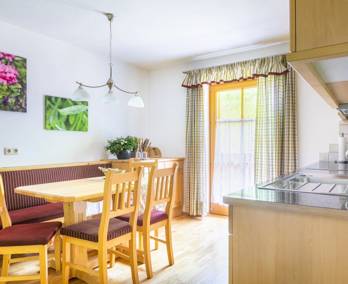 The farmhouse features a kitchen and dining area with a wooden table, bench seating, and chairs, a kitchen counter with a sink and stovetop, and a glass door providing outdoor access.