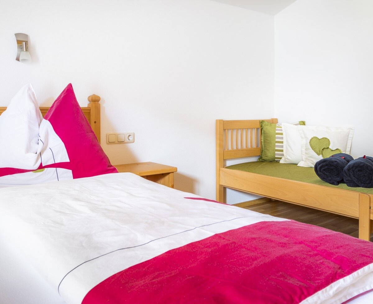 Farmhouse bedroom featuring a bed with red and white bedding, a wooden bedside table, and a daybed with green cushions and rolled towels.
