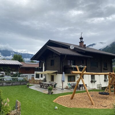 The farmhouse with a garden, patio, and children's playground against a mountain backdrop.