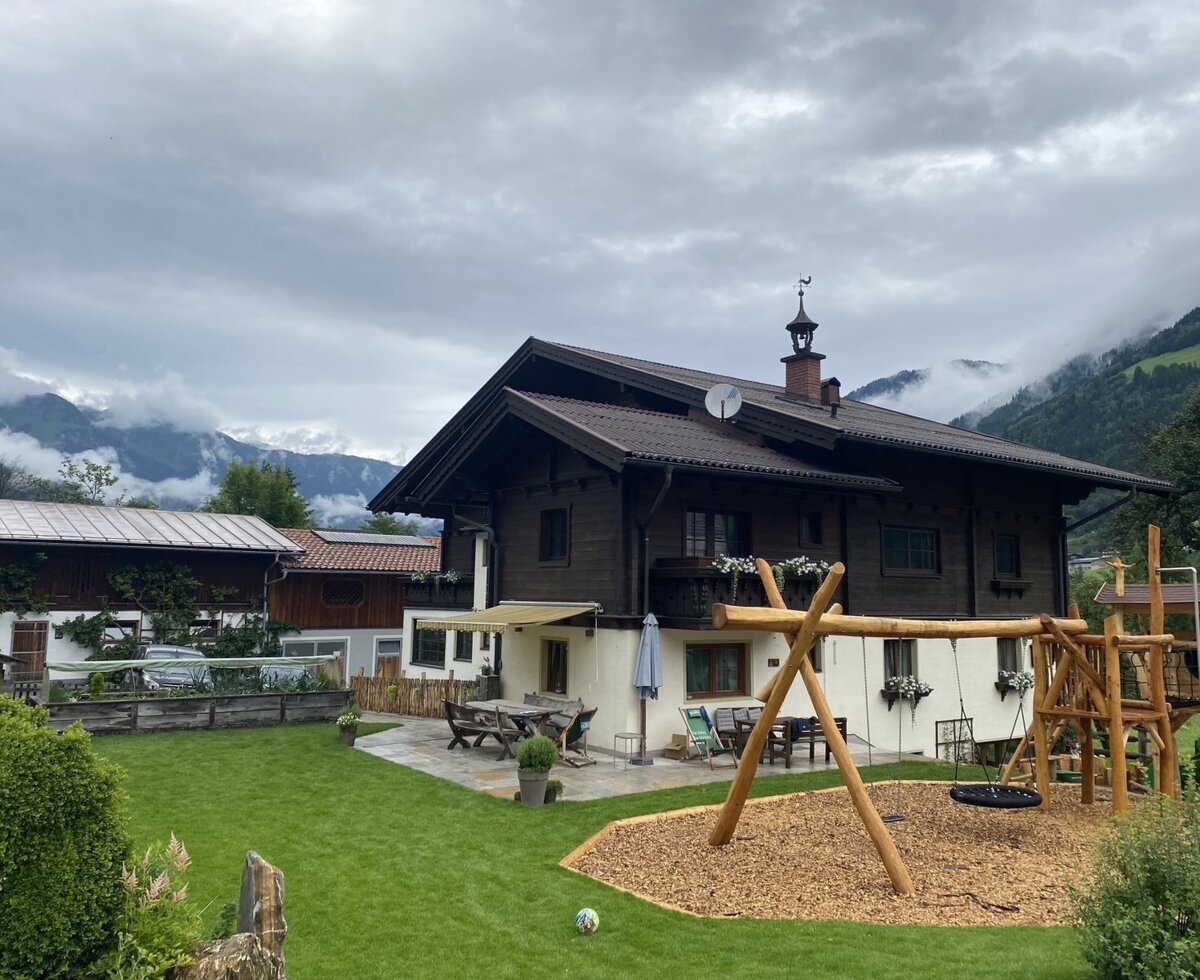 The farmhouse with a garden, patio, and children's playground against a mountain backdrop.