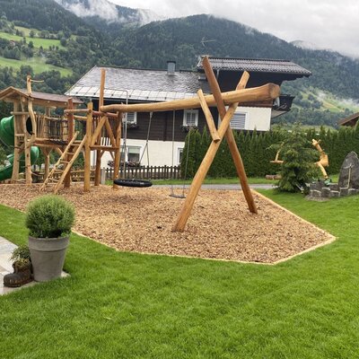 The farmhouse playground featuring a wooden play tower, slide, swing, and wood chip ground.