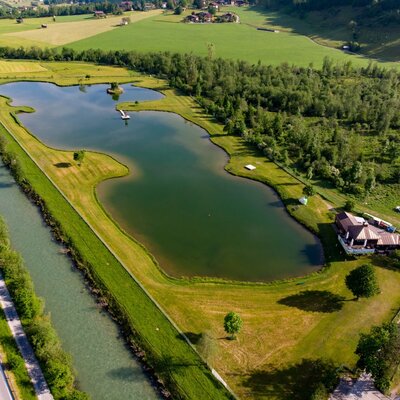 The swimming lake, surrounded by green spaces and trees, with a building on the shore and an adjacent canal.