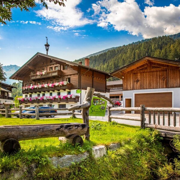 The farmhouse with a traditional wooden facade, balconies adorned with flowers, and an outbuilding, surrounded by green meadows and mountains.