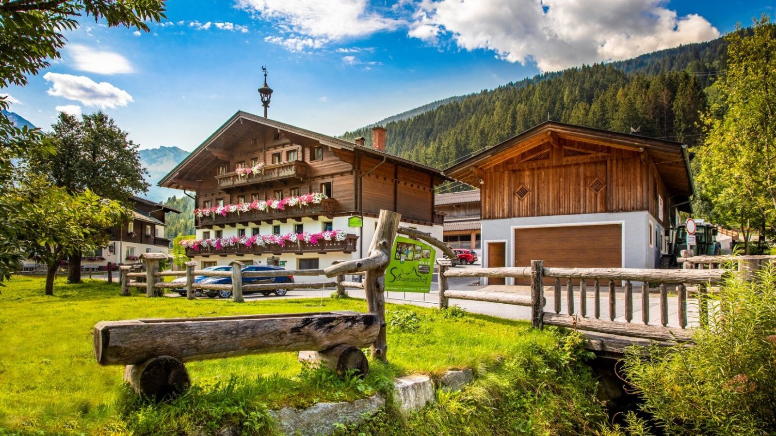 The farmhouse with a traditional wooden facade, balconies adorned with flowers, and an outbuilding, surrounded by green meadows and mountains.