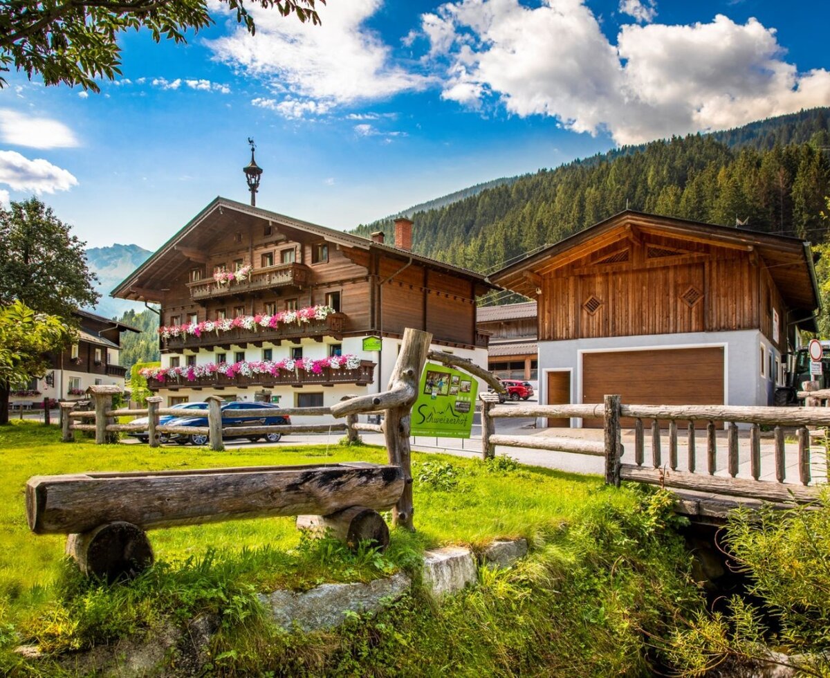 The farmhouse with a traditional wooden facade, balconies adorned with flowers, and an outbuilding, surrounded by green meadows and mountains.