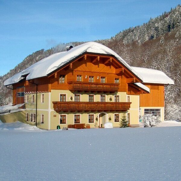 The farm house exterior in winter, featuring a snow-covered gabled roof, wooden balconies, and surrounding snowy fields and mountains.