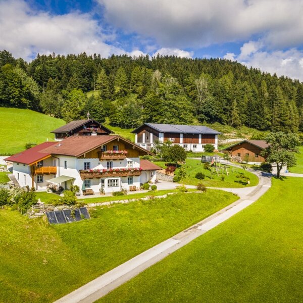 The Grubsteighof Farm House, situated on green hills backed by a forest, features multiple buildings and a children's playground.