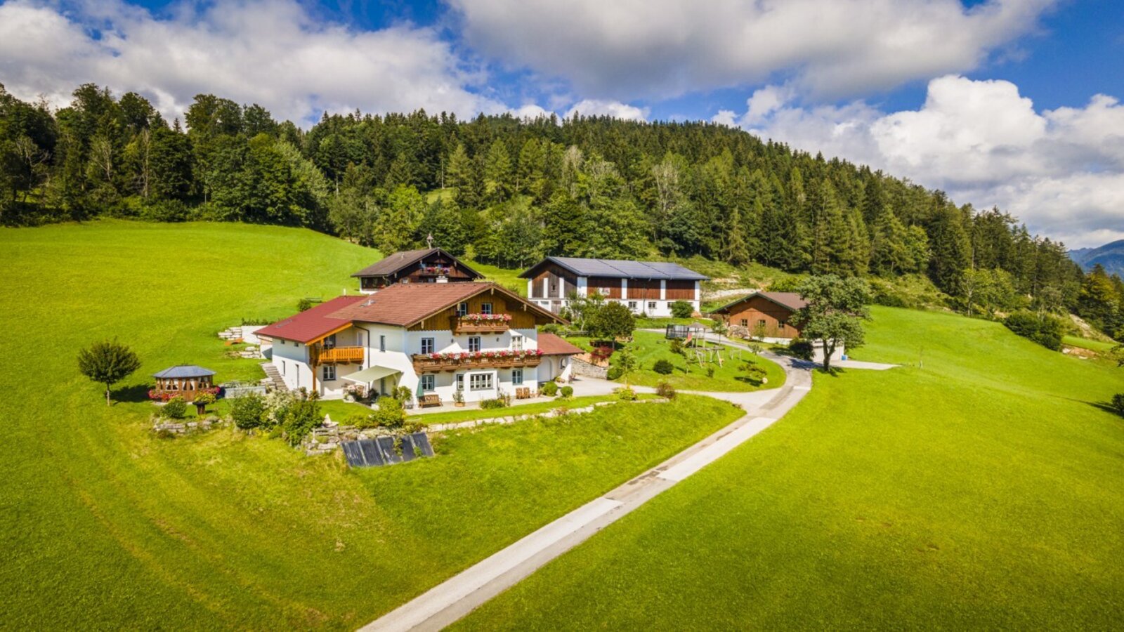 The Grubsteighof Farm House, situated on green hills backed by a forest, features multiple buildings and a children's playground.