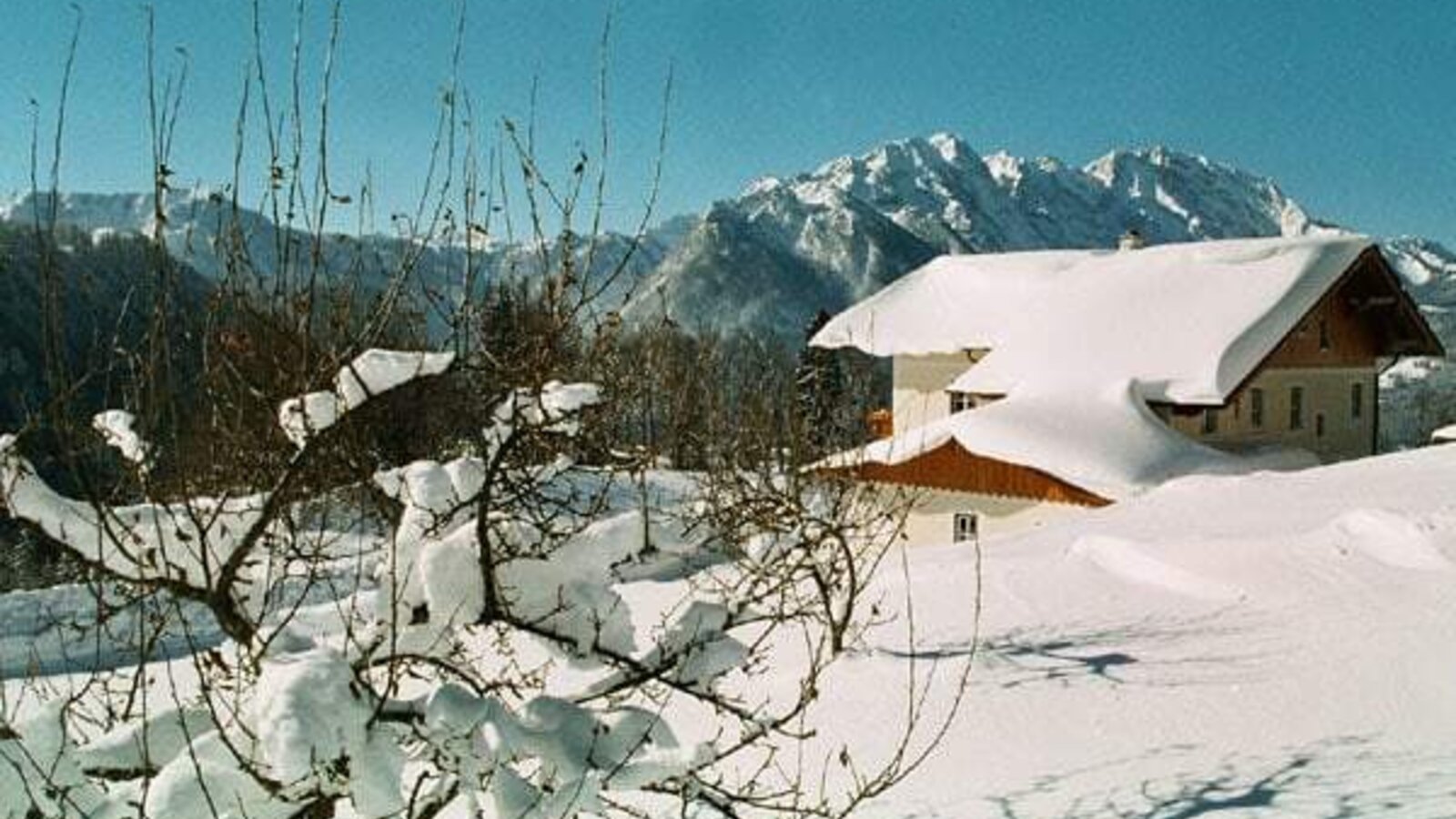 The Farm House exterior in winter, with its snow-covered roof and surrounding grounds, set against a backdrop of mountains.
