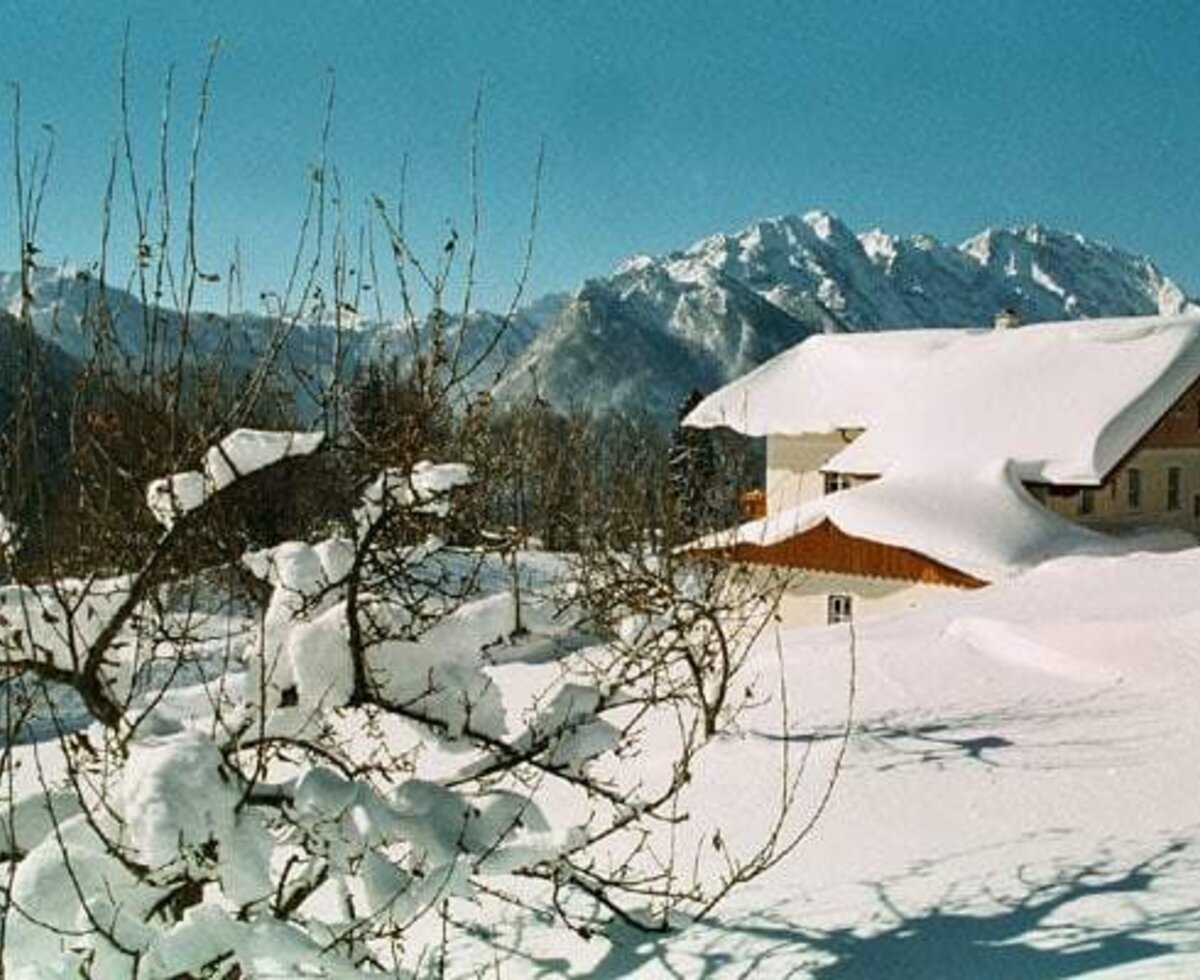 The Farm House exterior in winter, with its snow-covered roof and surrounding grounds, set against a backdrop of mountains.